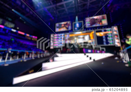 Blurred background of an esports event - Big illuminated main stage of a computer games tournament located on a big stadium. Tribunes are full of video games fans. Blurred background of an esports event - Big illuminated main stage of a computer games tournament located on a big stadium. Tribunes are full of video games fans. 65204893