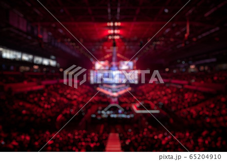 Blurred background of an esports event - Big illuminated main stage of a computer games tournament located on a big stadium. Tribunes are full of video games fans. Blurred background of an esports event - Big illuminated main stage of a computer games tournament located on a big stadium. Tribunes are full of video games fans. 65204910