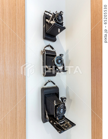 Vintage cameras with a black, leather case hanging in a corner on a white wall. Vintage cameras with a black, leather case hanging in a corner on a white wall. 65205530