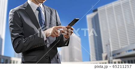 高層ビル街でスマートフォンを操作する若いビジネスマンのイメージ素材 65211257