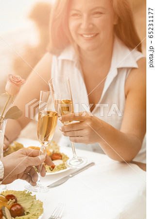 Happy woman smiles holding glass of white wine. Mature couple have event outdoors at beach cafe. Selective focus on human hands with glasses of wine or champagne in foreground. Toned image 65231177