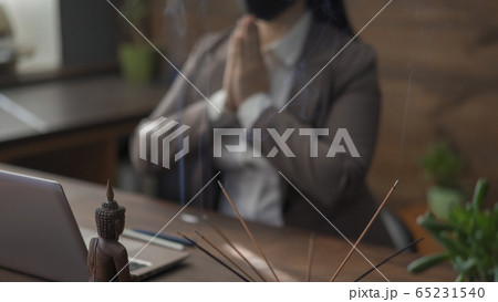 Businesswoman woman praying on a blurred background. Abstract picture of a modern office in oriental style, selective focus on incense sticks and a Buddha figurine in the foreground Businesswoman woman praying on a blurred background. Abstract picture of a modern office in oriental style, selective focus on incense sticks and a Buddha figurine in the foreground 65231540