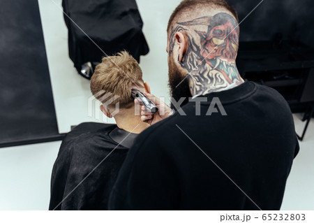Male haircut with electric razor. Tattooed Barber makes haircut for client at the barber shop by using hairclipper. Man hairdressing with electric shaver. 65232803