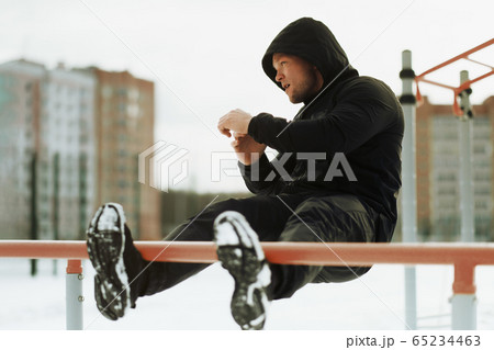Caucasian young man is engaged in fitness and sports in winter outdoors. Street workout. Athlete trains on a street turnstile in black sportswear. Caucasian young man is engaged in fitness and sports in winter outdoors. Street workout. Athlete trains on a street turnstile in black sportswear. 65234463