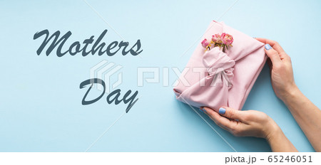 Women's hands are holding a holiday gift wrapping fabric Furoshiki style on a pink background lettering Mother's Day. 65246051