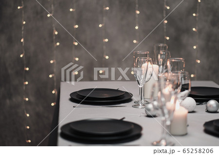 A table set in the restaurant. Serving a table. Grey artistic background. Black matte utensils, glass glasses. White candles and decor. Festive yellow lights garland in the background. 65258206