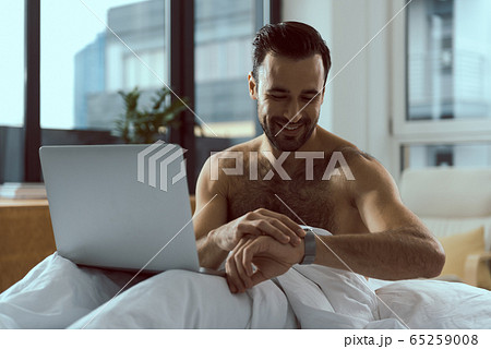 Happy man with notebook looking at watch in bed Happy man with notebook looking at watch in bed 65259008
