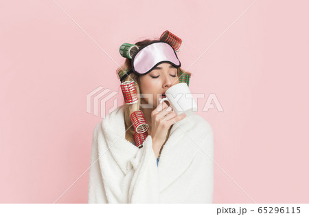 Sleepy woman in curlers and sleep mask wrapped in blanket and drinks coffee from cup 65296115