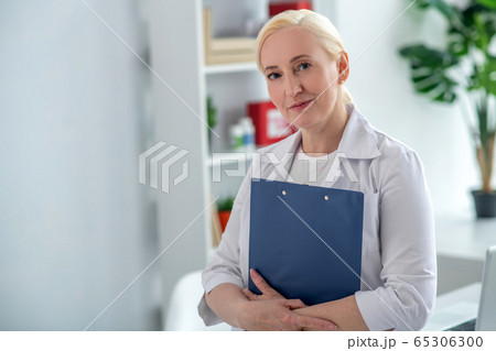 Blonde middle-aged doctor holding documents and looking positively Blonde middle-aged doctor holding documents and looking positively 65306300