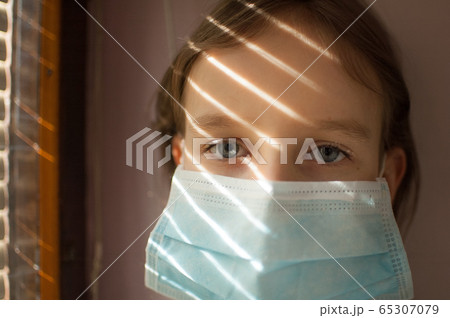 Coronavirus Covid-19 outbreak. Little blonde girl with blue eyes wearing disposable mask for protection of virus sitting near the window with blinds. Personal protective equipment Coronavirus Covid-19 outbreak. Little blonde girl with blue eyes wearing disposable mask for protection of virus sitting near the window with blinds. Personal protective equipment 65307079