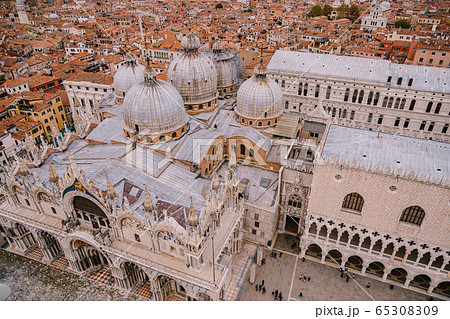 Top view of St. Mark's Cathedral in St. Mark's Square, with a large brick tower. Against the background of tiled orange roofs of the old city of Venice in Italy. Top view of St. Mark's Cathedral in St. Mark's Square, with a large brick tower. Against the background of tiled orange roofs of the old city of Venice in Italy. 65308309