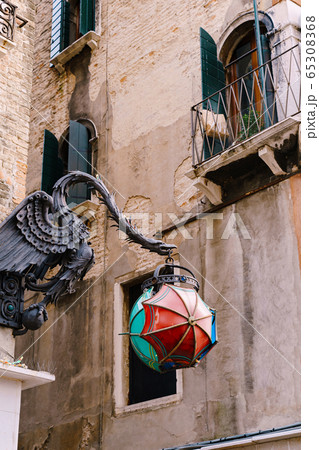 An unusual street lamp in the shape of The Maforio Dragon on the corner of the building, with a ball of three umbrellas - blue, green and red, on the streets of Venice, Italy, near the Rialto bridge. An unusual street lamp in the shape of The Maforio Dragon on the corner of the building, with a ball of three umbrellas - blue, green and red, on the streets of Venice, Italy, near the Rialto bridge. 65308368