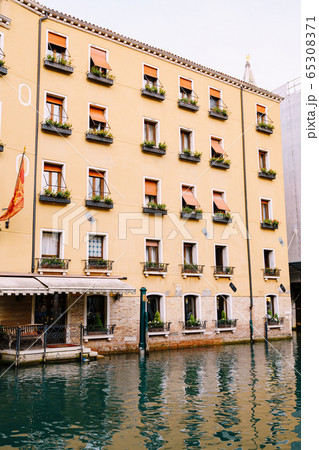 Yellow five-story building in Venice, Italy. The main entrance of the building goes into the water, at the entrance there is a sofa, on horseback there are flower pots and orange shutters from the sun 65308371