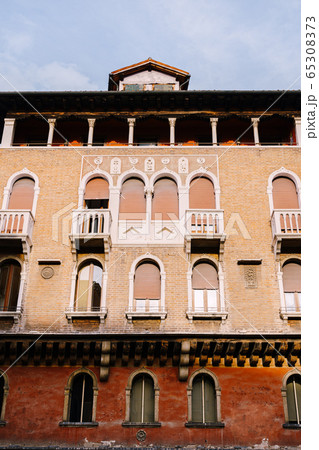 A four-story brown brick building in Venice, Italy. Narrow arched windows, with small balconies with columns, brown plastic sunblinds. Classical venetian architecture in the facade of the building. 65308373