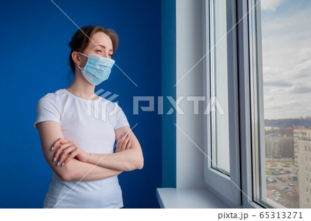 Pensive woman wearing medical face mask and looking out of window in blue room 65313271