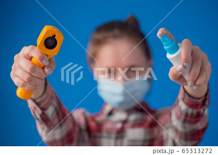 Woman holding pyrometer and spraying antiseptic toward camera against blue wall 65313272
