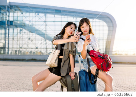 Two charming young dark haired asian women in casual summer clothes using their smartphones for social networks or checking flight time, sitting on the suitcases in front of airport building Two charming young dark haired asian women in casual summer clothes using their smartphones for social networks or checking flight time, sitting on the suitcases in front of airport building 65336662