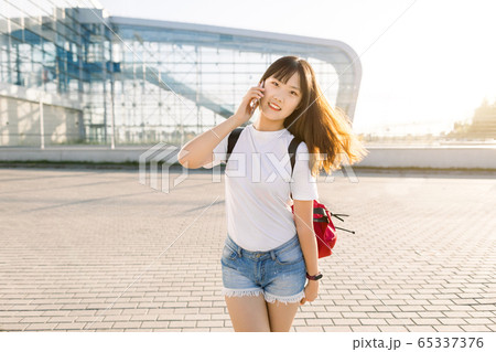 Pretty cheerful 25-aged asian brunette girl in jeans shorts and white t-shirt enjoying her conversation with friend on smartphone in front of modern airport building. Travel and technologies concept Pretty cheerful 25-aged asian brunette girl in jeans shorts and white t-shirt enjoying her conversation with friend on smartphone in front of modern airport building. Travel and technologies concept 65337376