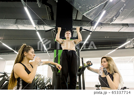 shot of a muscular Caucasian male athlete doing pull ups at the gym. Strong energetic sportsman pulling up on a bar at sport studio, showing workout for two pretty girls shot of a muscular Caucasian male athlete doing pull ups at the gym. Strong energetic sportsman pulling up on a bar at sport studio, showing workout for two pretty girls 65337714
