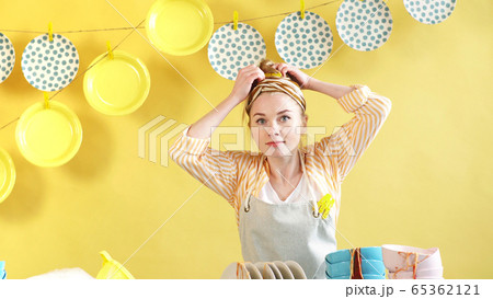 young cheerful woman put on an apron and collects her hair in a bun, before starting homework young cheerful woman put on an apron and collects her hair in a bun, before starting homework 65362121