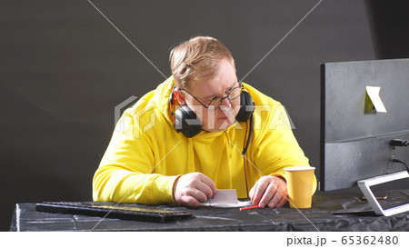 Plump man in a yellow jacket is sitting at a computer on an isolated black background 65362480