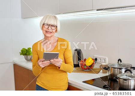 Retirement. Senior woman in glasses standing at kitchen browsing internet looking for recipe on digital tablet smiling happy Retirement. Senior woman in glasses standing at kitchen browsing internet looking for recipe on digital tablet smiling happy 65363501