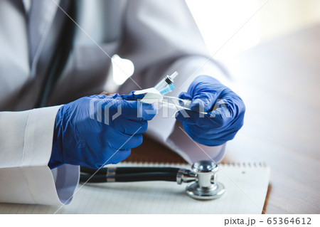 Close up of doctors hands wearing blue protective gloves with stethoscope and syringe on wooden table background Close up of doctors hands wearing blue protective gloves with stethoscope and syringe on wooden table background 65364612