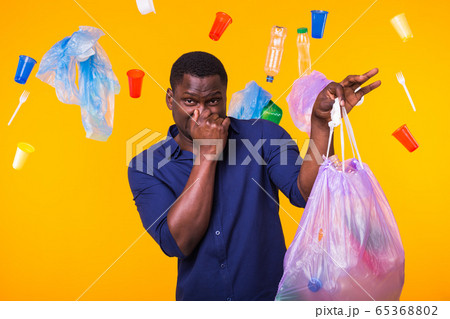 Environmental pollution, plastic recycling problem and waste disposal concept - angry african american man holding garbage bag on yellow background. He is feel smell of trash. 65368802