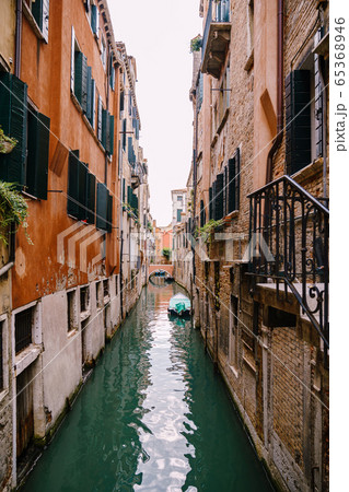 Beautiful narrow canal in Venice, Italy. Between two buildings. A small bridge in distance. Moored boat against the wall of the house. The left building is orange, the right one is made of bare brick. Beautiful narrow canal in Venice, Italy. Between two buildings. A small bridge in distance. Moored boat against the wall of the house. The left building is orange, the right one is made of bare brick. 65368946