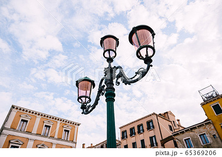 An ancient triple metal street lamp, with pink plafonds against the sky and buildings of Venice, Italy. Vintage street lighting, elegant pillar design. An ancient triple metal street lamp, with pink plafonds against the sky and buildings of Venice, Italy. Vintage street lighting, elegant pillar design. 65369206