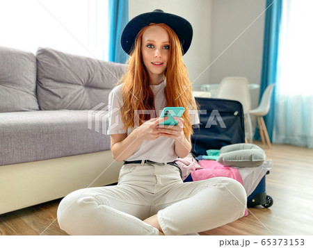 Pretty woman with red hair in a black hat is sitting at home near a suitcase with phones in her hands, waiting for a taxi to the airport. The concept of travel 65373153