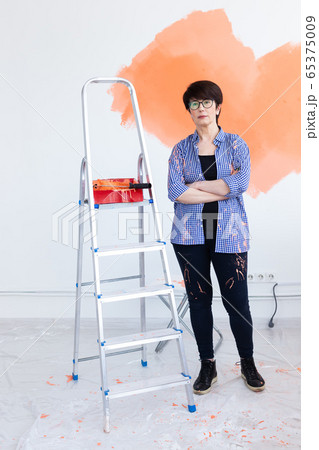 Pretty smiling middle-aged woman painting interior wall of home with paint roller. Redecoration, renovation, apartment repair and refreshment concept. 65375009