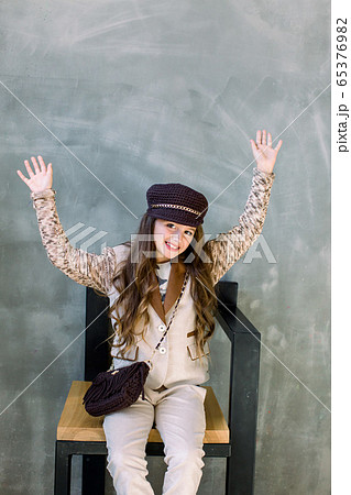 portrait of beautiful delighted little lady in a light suit, a golden sneakers and a hat is sitting on the armchair waving her hands portrait of beautiful delighted little lady in a light suit, a golden sneakers and a hat is sitting on the armchair waving her hands 65376982
