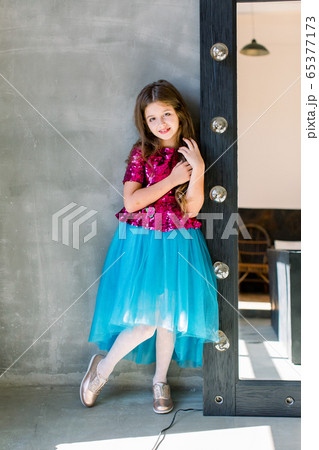 Beautiful cute little girl in a pink shiny blouse and a blue skirt on a grey background standing near mirror. Beautiful cute little girl in a pink shiny blouse and a blue skirt on a grey background standing near mirror. 65377173