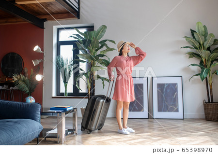 Dark-haired female standing with her suitcase, touching her straw hat with one hand 65398970