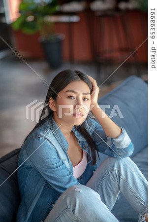 Upset dark-haired female sitting with folded knees on sofa, thinking, resting her head on one hand Upset dark-haired female sitting with folded knees on sofa, thinking, resting her head on one hand 65398991