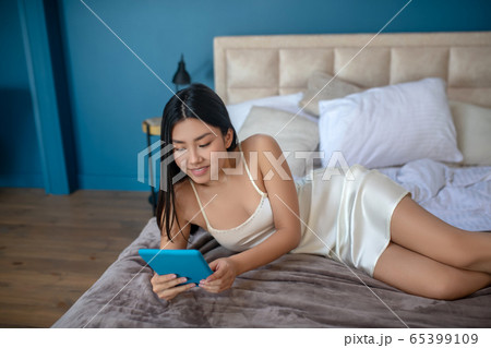 Young brunette enjoying her morning, lying on her side, checking something on her tablet, smiling 65399109