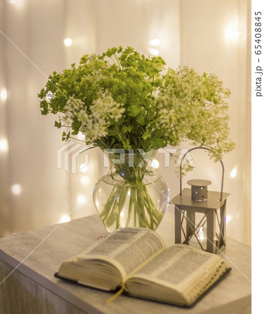 Composition: an open Bible, a bouquet of wildflowers in a vase and a decorative flashlight with a candle on the nightstand against the background of burning lights. Composition: an open Bible, a bouquet of wildflowers in a vase and a decorative flashlight with a candle on the nightstand against the background of burning lights. 65408845