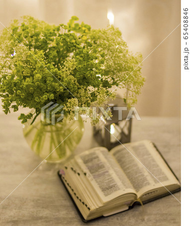 Composition: an open Bible, a bouquet of wildflowers in a vase and a decorative flashlight with a candle on the nightstand against the background of burning lights. 65408846