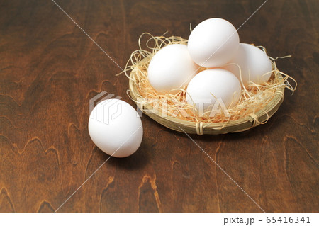 わらを敷いた小さいザル盛り白い鶏卵四個、茶色い木目バック 65416341