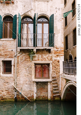 Close-ups of building facades in Venice, Italy. Beautiful Venetian windows with open wooden green shutters. An old red brick house next to a bridge over a small narrow canal. 65426514