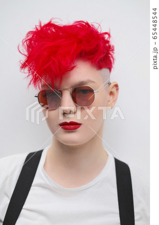 Portrait of a cool fashionable modern young girl. A short haircut with shaved temple. Dyed bright red hair. Red lipstick. Studio photo on white background. Suspenders and checkered pants. 65448544