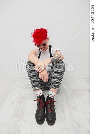 Portrait of a cool fashionable modern young girl. A short haircut with shaved temple. Dyed bright red hair. Red lipstick. Studio photo on white background. Suspenders and checkered pants. 65448555