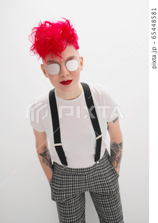 Portrait of a cool fashionable modern young girl. A short haircut with shaved temple. Dyed bright red hair. Red lipstick. Studio photo on white background. Suspenders and checkered pants. 65448581