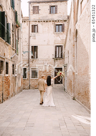 Italy wedding in Venice. The bride and groom walk along the deserted streets of the city. Newlyweds are walking in a dead end alley on the background of brick buildings. Italy wedding in Venice. The bride and groom walk along the deserted streets of the city. Newlyweds are walking in a dead end alley on the background of brick buildings. 65451322