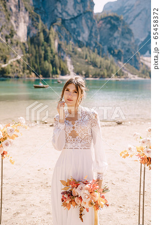 Beautiful bride in a white dress with sleeves and lace, with a yellow autumn bouquet on background of the arch for ceremony, at Lago di Braies in Italy. Destination wedding in Europe, on Braies lake. 65458372