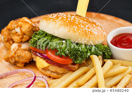 Combo set of fast food. Juicy hamburger, French fries, chicken legs and sauce. Served wooden board and cutlery on a black background. Fast food restaurant. Food delivery. 65480240