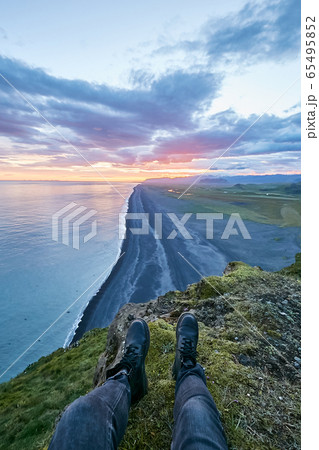 Traveler sitting on the cape above amazing black sand beach in Iceland and watching sunset or sunrise with soft pink clouds. Legs hanging from the cliff 65495852