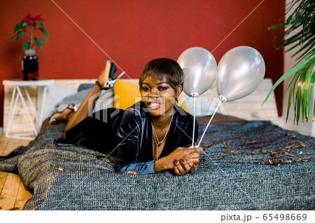 Happy dark-skinned girl in a dark evening dress is lying on the bed after the birthday party. 65498669