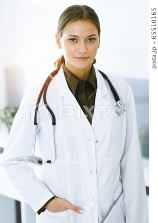 Woman-doctor standing and looking at camera in sunny clinic. Perfect medical service. Medicine and healthcare Woman-doctor standing and looking at camera in sunny clinic. Perfect medical service. Medicine and healthcare 65510165